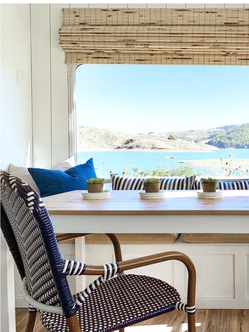 Interior shot of an RV, with a dining room table, chair, and bench filled with pillows for a comfortable seating area. The window behind the bench looks out to blue water and green trees on a sunny day.