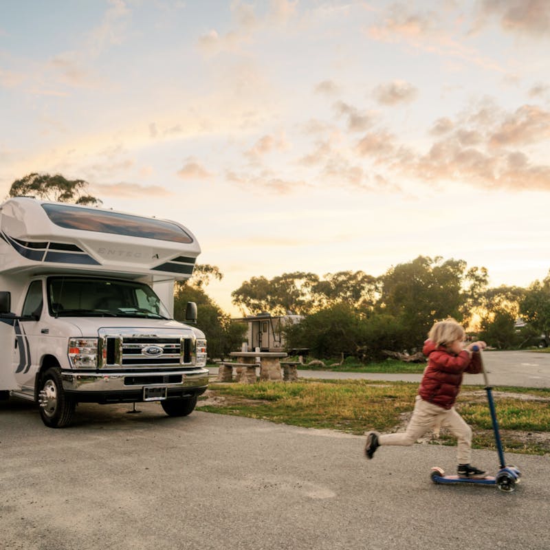 Little boy rides scooter down pavement with class c motorhome in background at sunset
