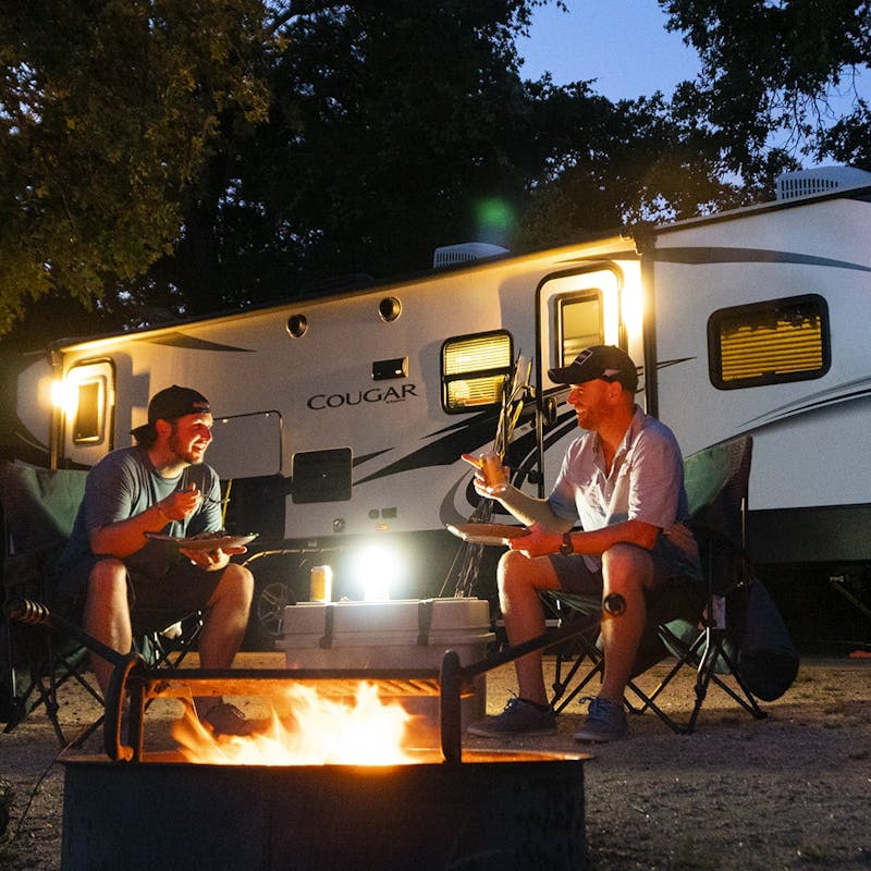 Two men sitting by a campfire, with an RV in the background, eating dinner and laughing