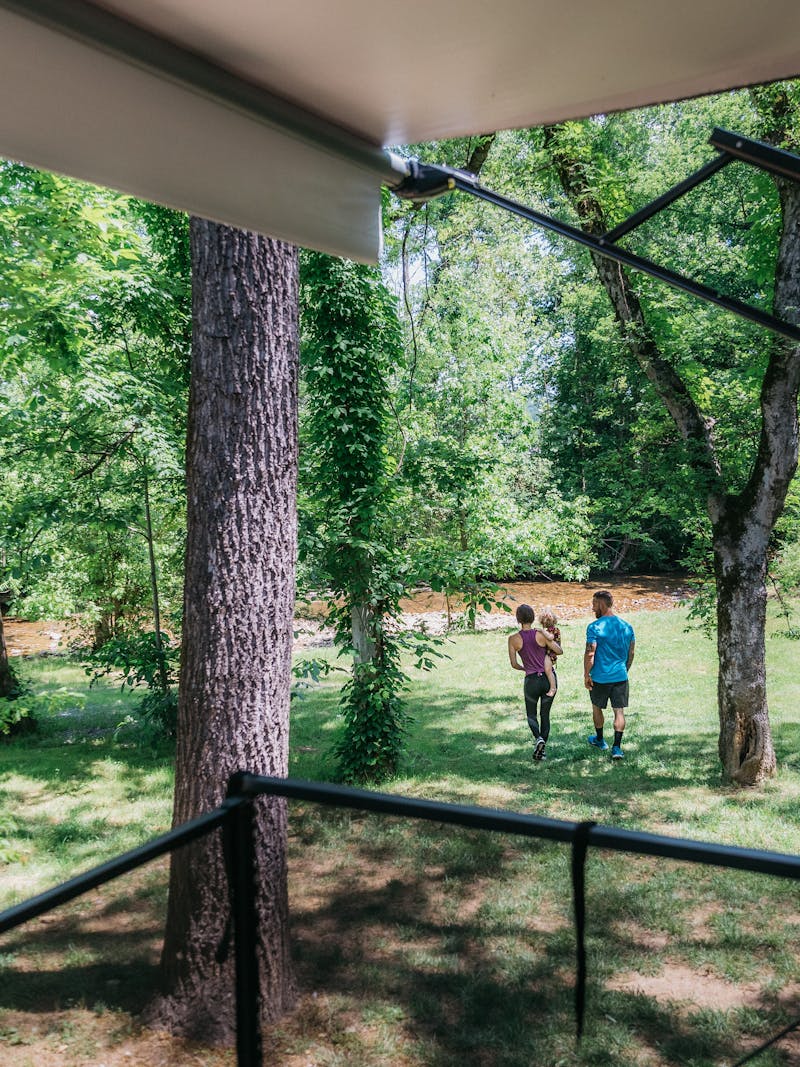 A young mom and dad walk their toddler daughter across a grassy lawn toward a stream.