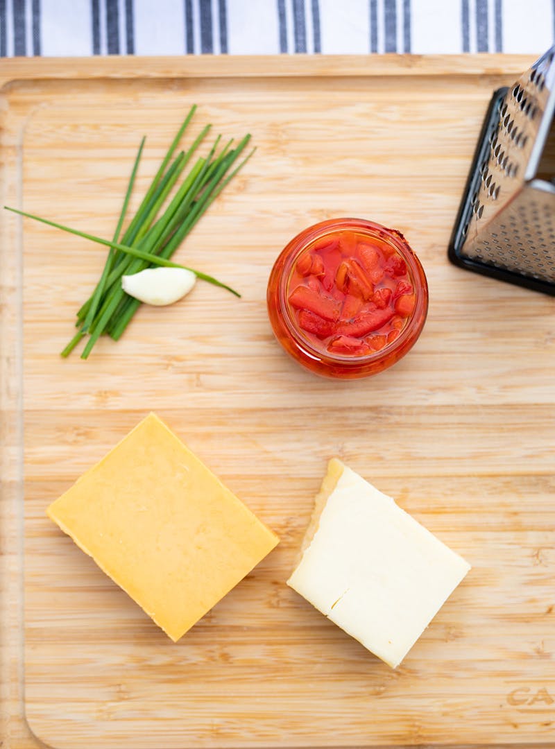 A cutting board with chives, garlic, cheese and a jar of pimentos on it.
