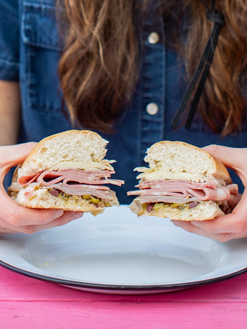 A woman holding a muffaletta sandwich up to show if off to the camera.