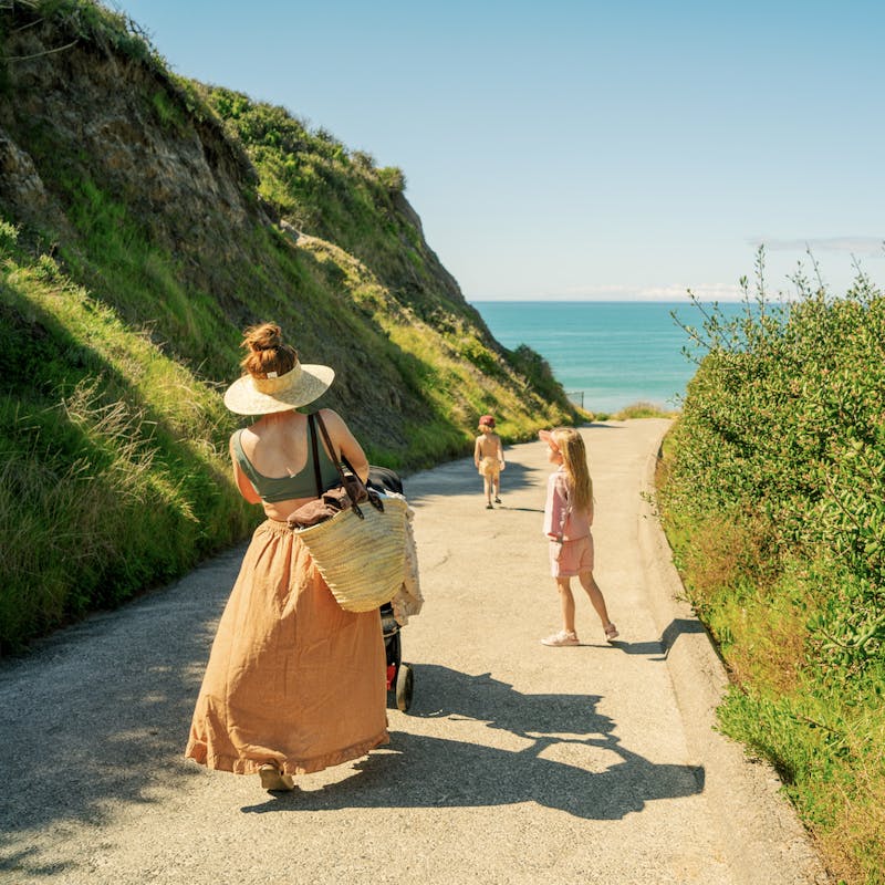 Mom in orange skirt pushes stroller down paved road with two kids and ocean in background