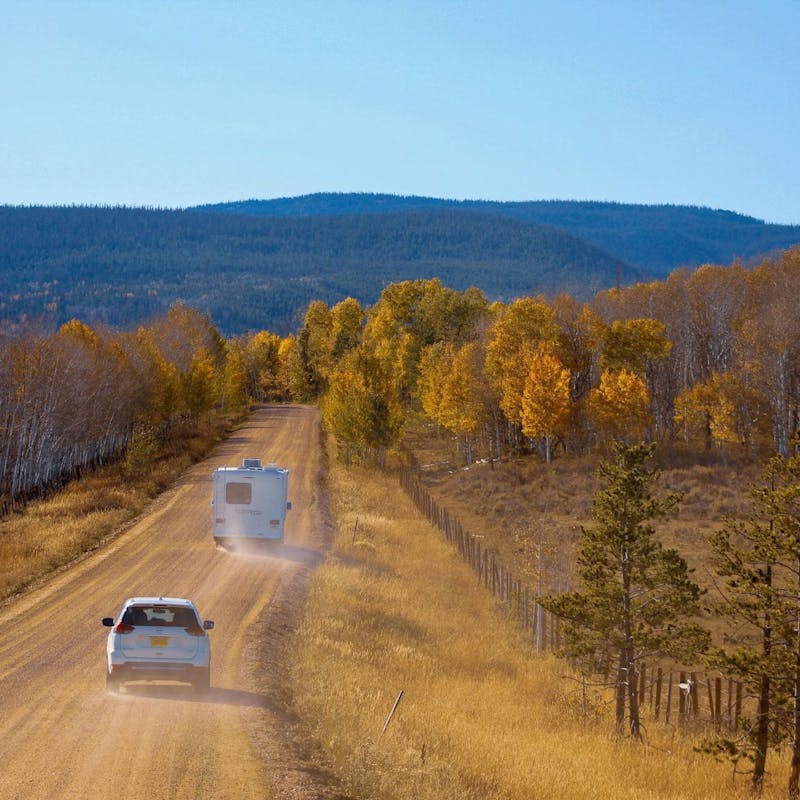 White car and Class C RV on dusty dirt road heading into forest of yellow trees