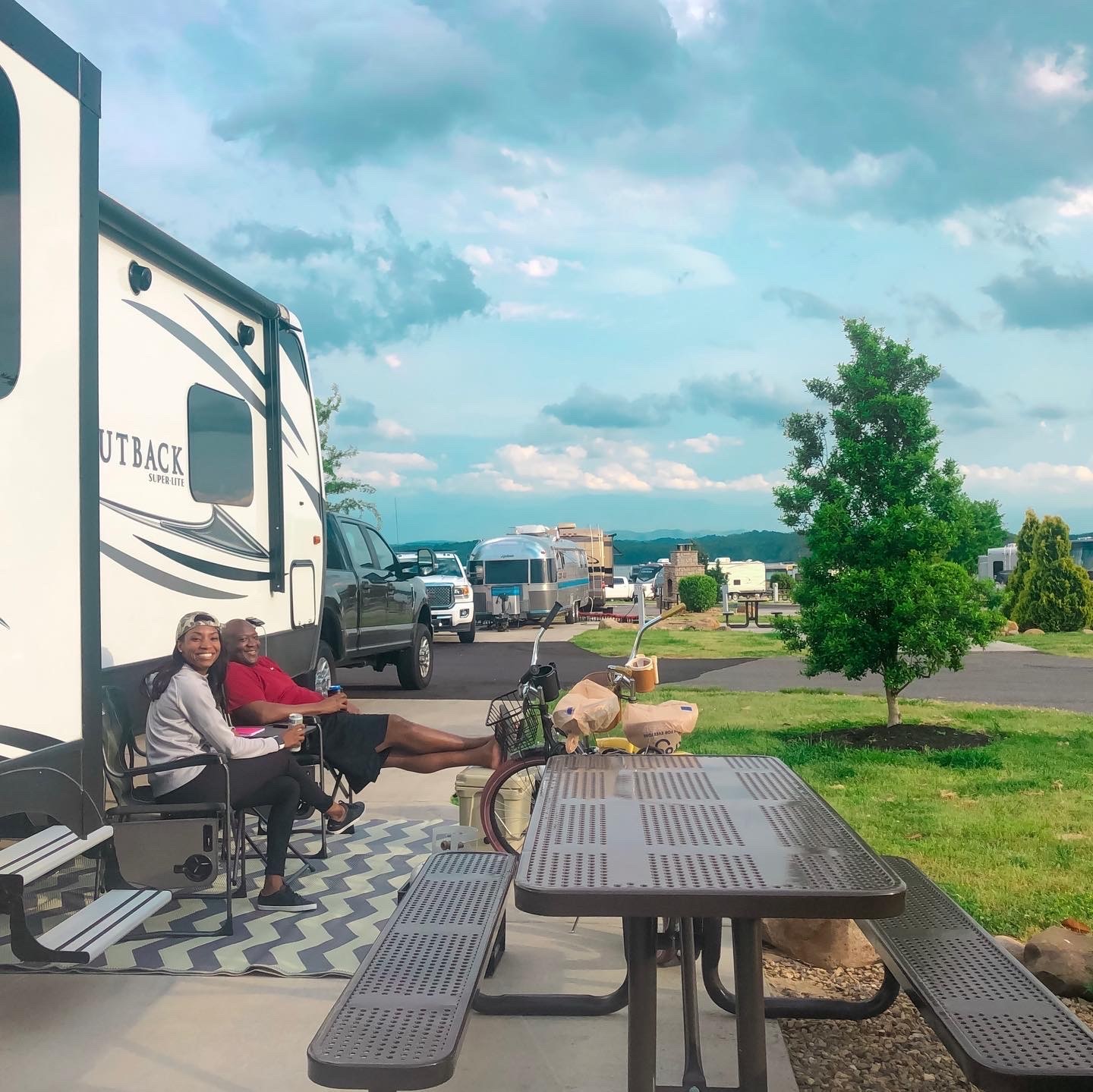 A couple relaxes outside their RV in camping chairs