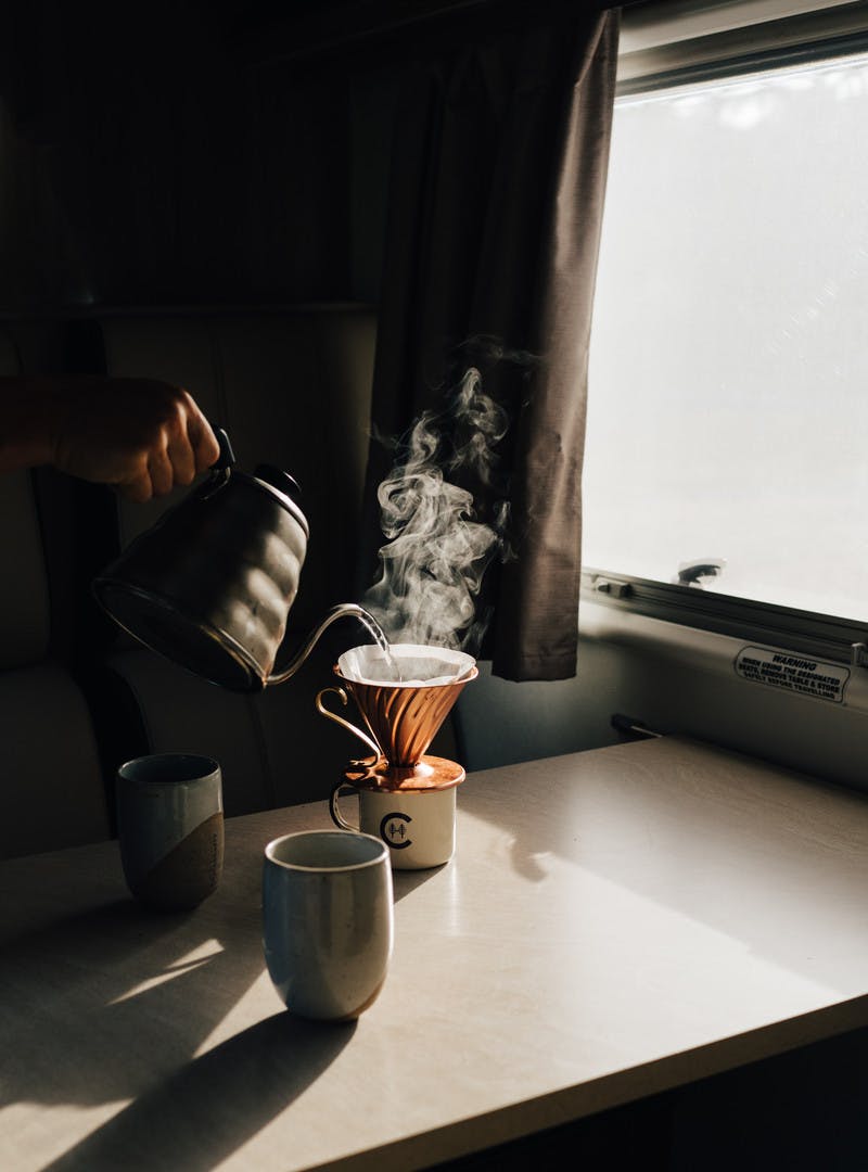 Next to a window, a shadowy shot of a hand pouring water out of a kettle over coffee grounds inside a Class C motorhome.