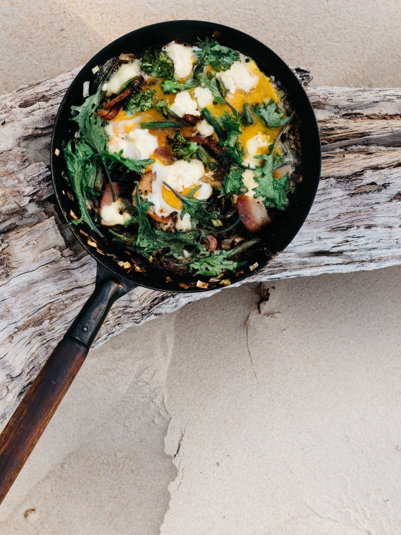 A cast iron skilled filled with cooked greens, bacon and eggs, balanced on a piece of driftwood on the beach. 