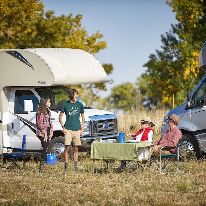 Two Class C RVs parked outside with people chatting in front of them