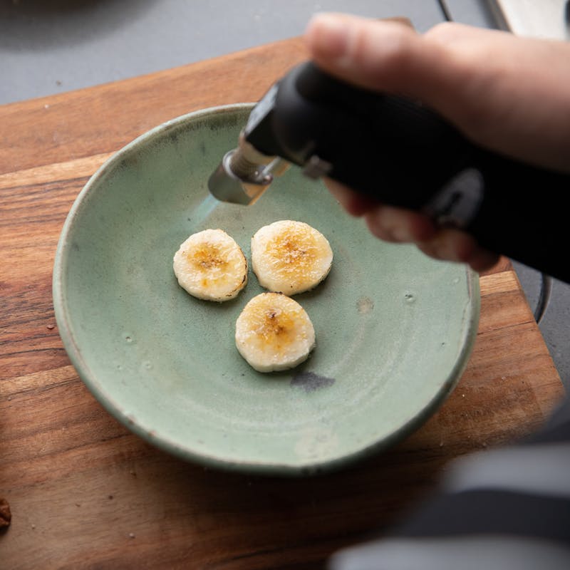 A hand using a kitchen blowtorch to caramelize the tops of banana slices.