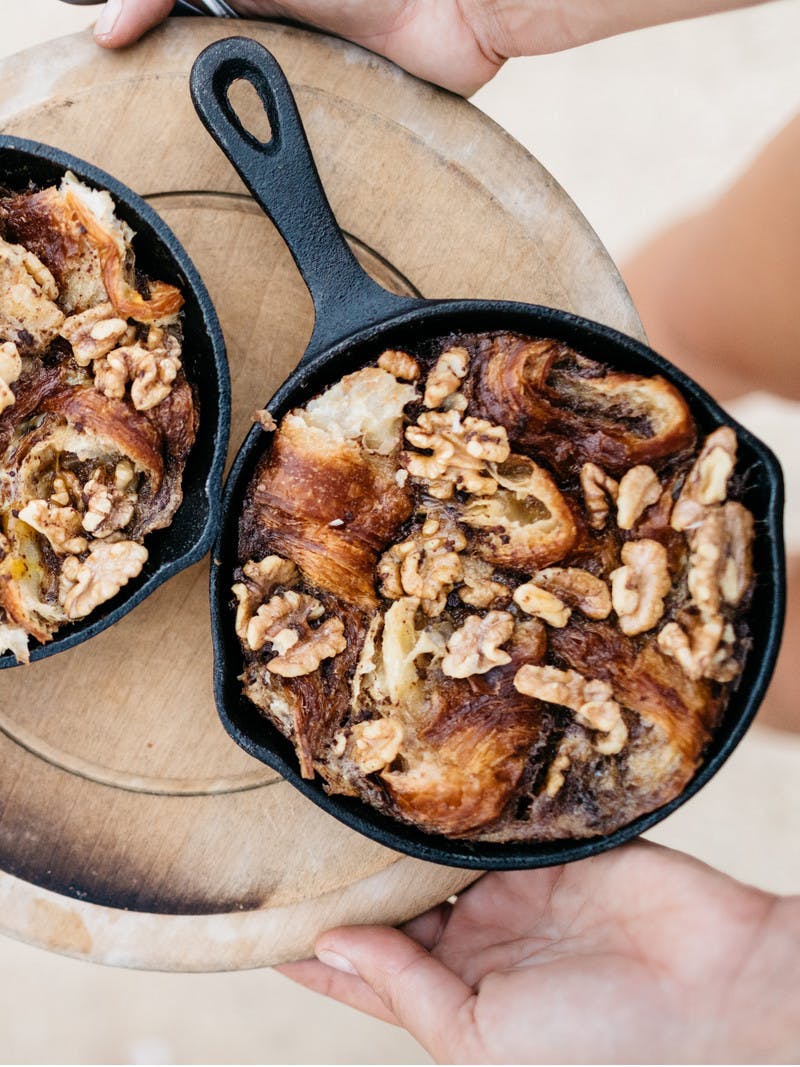 Two mini cast iron skillets on a tray, holding warm caramelized maple walnut croissant pudding.