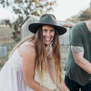 Sarah Glover, in a white sun dress and a felt hat, cooking over grill and smiling at the camera.