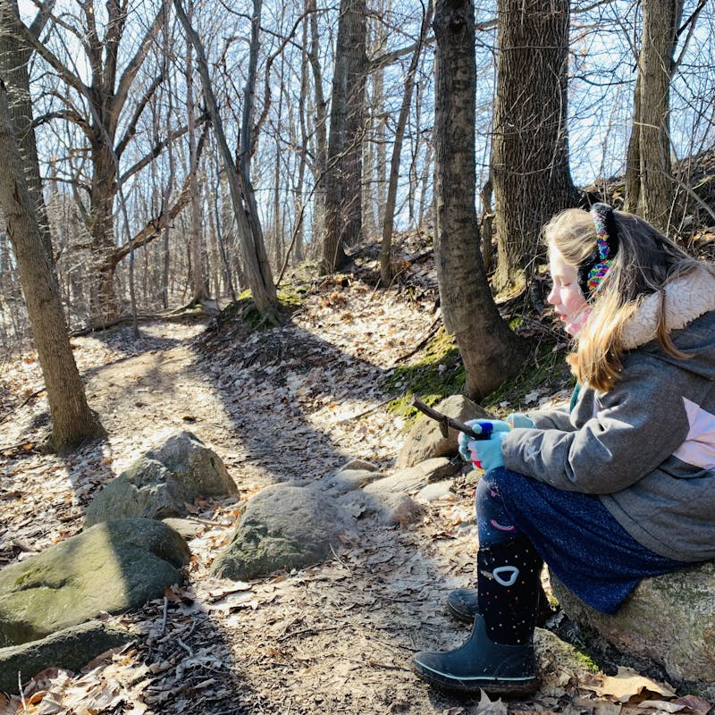 Young girl wearing snow jacket and earmuffs sits on a long and admires a stick