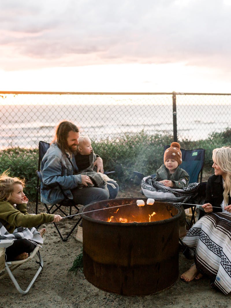 Amber Thrane and her family roasting marshmallows over a fire next to an Airstream Travel Trailer RV, with the ocean in the background.