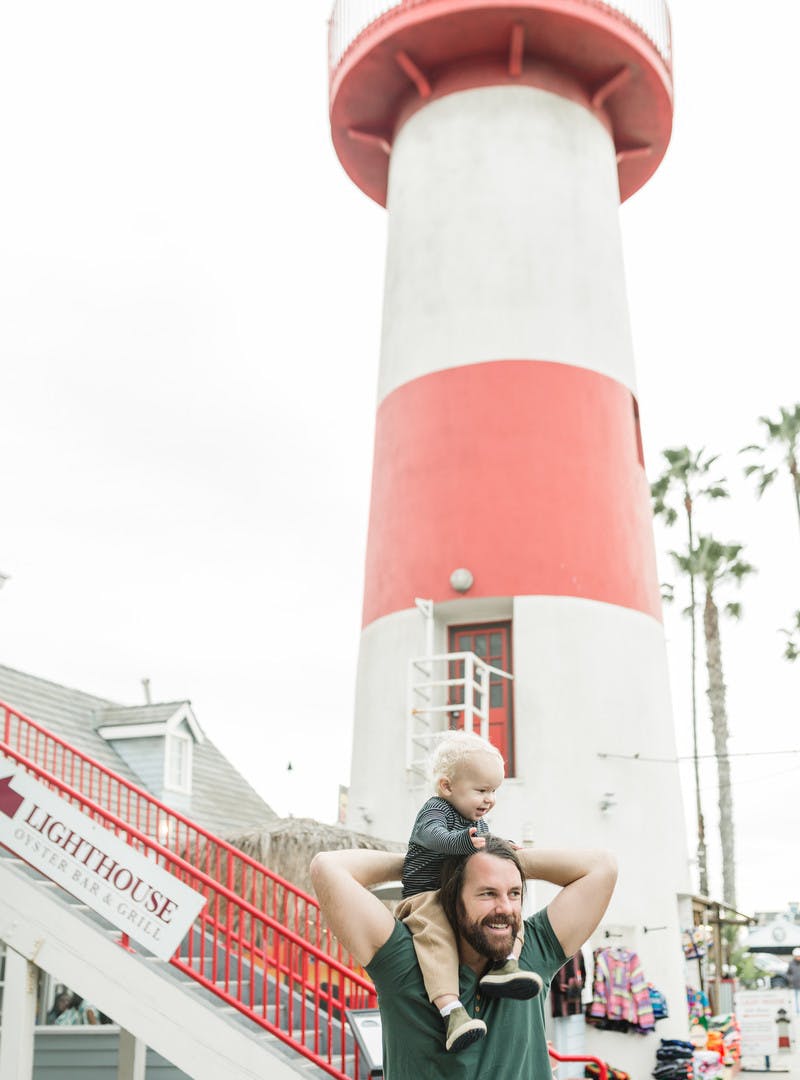 Man holds young child on his shoulders in front of a red and white lighthouse.