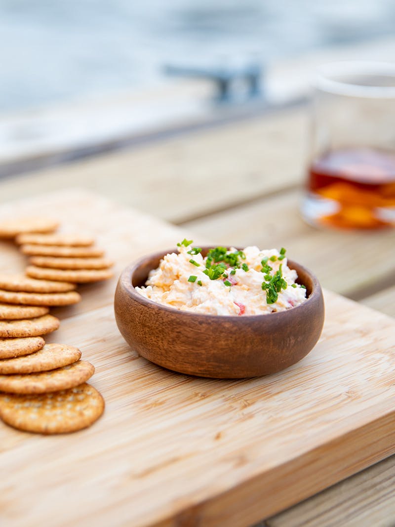 A cheese board with crackers, salami, pickled vegetables and a bowl of pimento cheese spread.