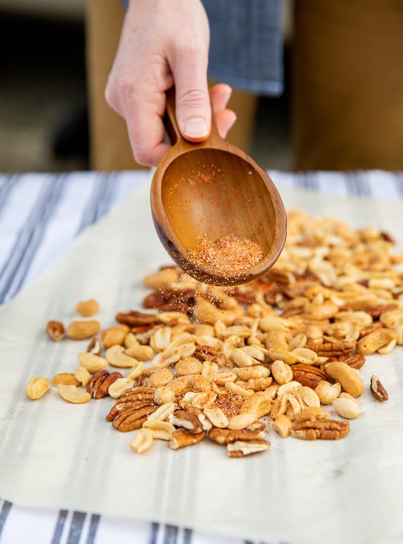 A hand holding a wooden spoon sprinkles spices over mixed nuts.