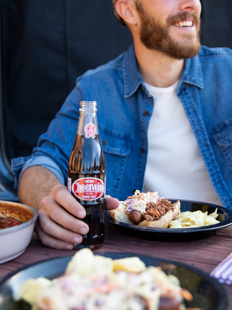 A man smiles holding a soda while sitting at a table and a Carolina Hot Dog plated in front of him.