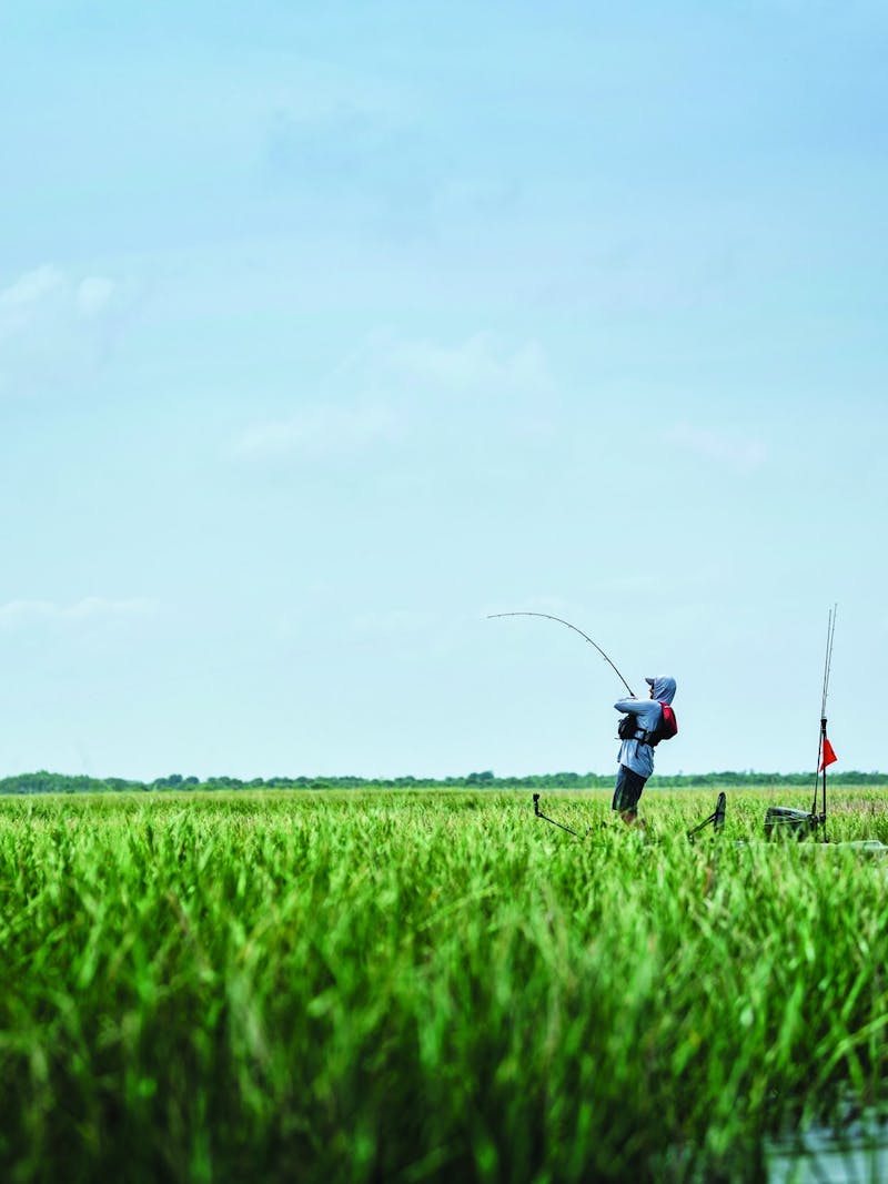 Kayak fisherman reels in fish standing among river grass