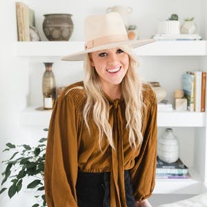Amber Thrane in a brown shirt, wearing a large cream colored hat, standing in front of white shelves, smiling at the camera.