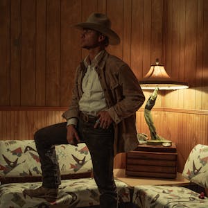 Man in a cowboy hats standing in wood paneled room, looking away from the camera, with leg up on a couch