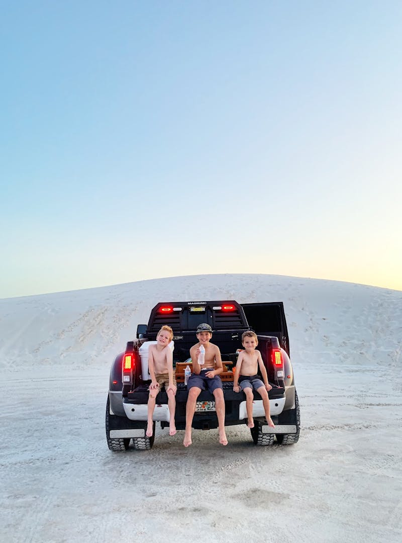 Three boys sitting on the open bed of a pickup truck while it's parked.