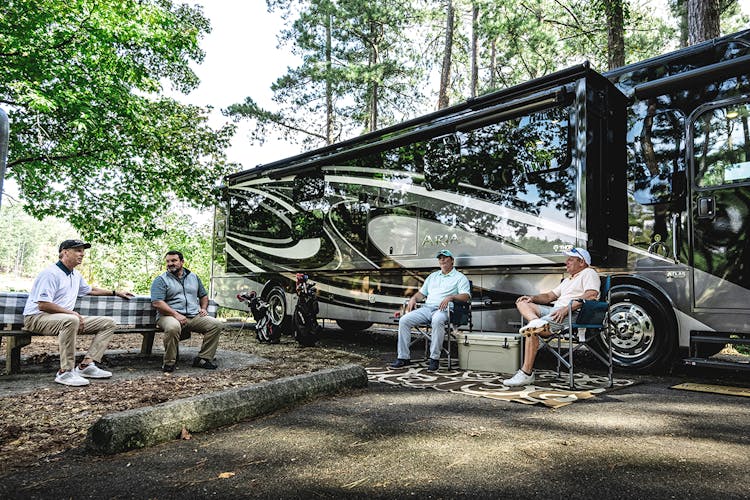 A Thor motor coach parked at a camp site, four men are sitting out front having a conversation while grilling food.
