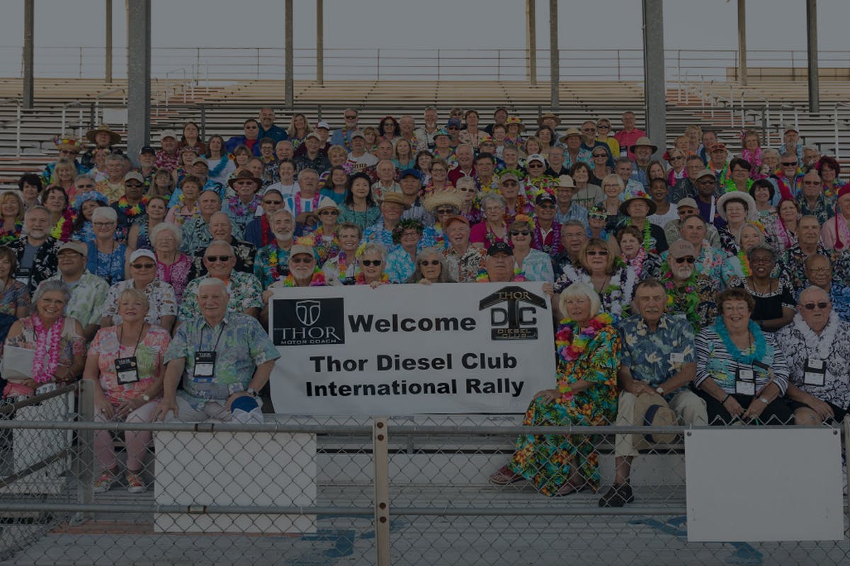 A group of motor coach owners sitting in bleachers together, in front of them is a large sign that says welcome Thor diesel club international rally.
