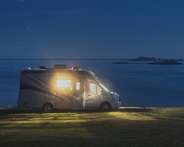 A motor home parked next to the shore at night with the light on illuminating its surroundings