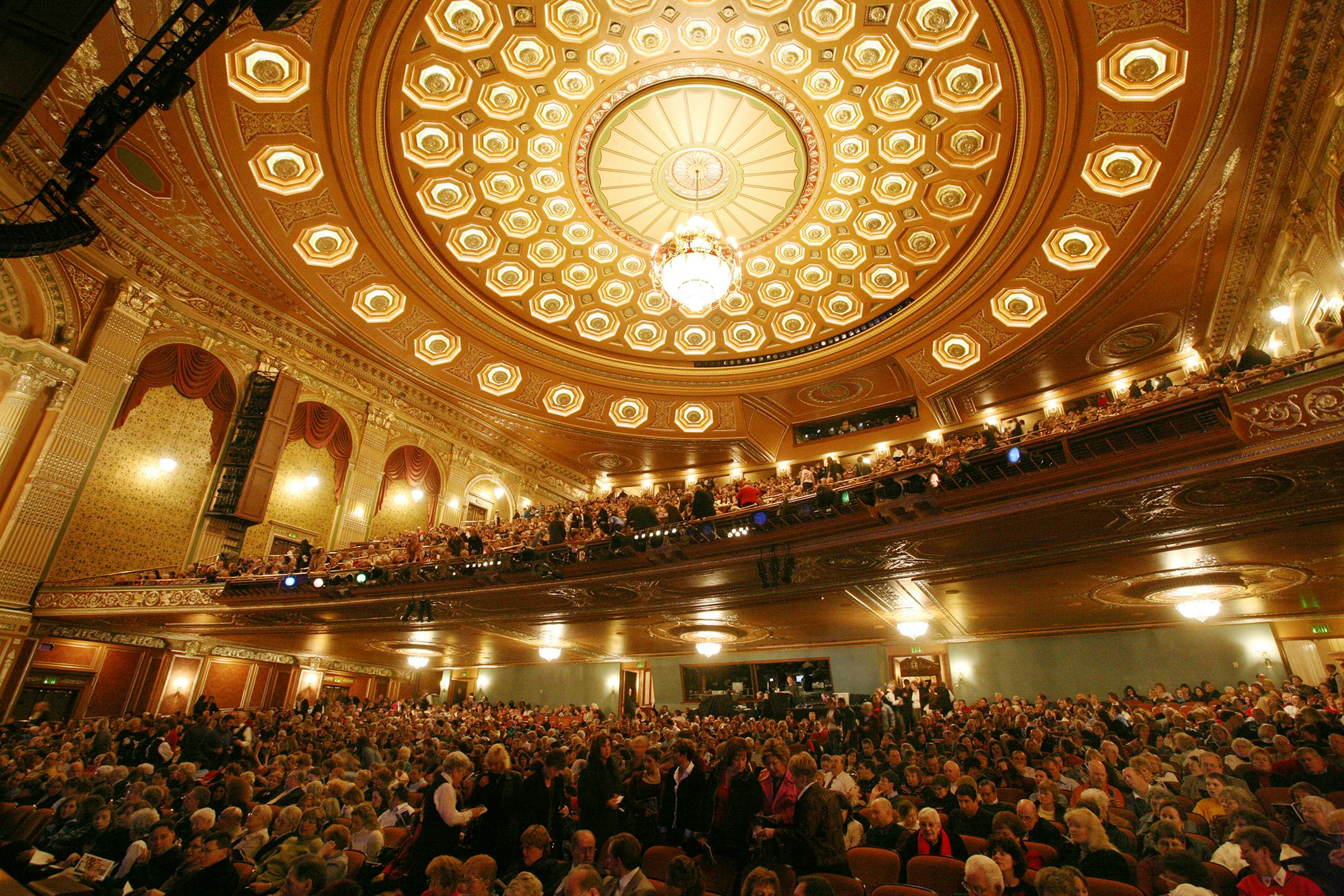 The ceiling of the Benedum Center concert hall, featuring ornate gold designs and a large, brightly-lit chandelier.