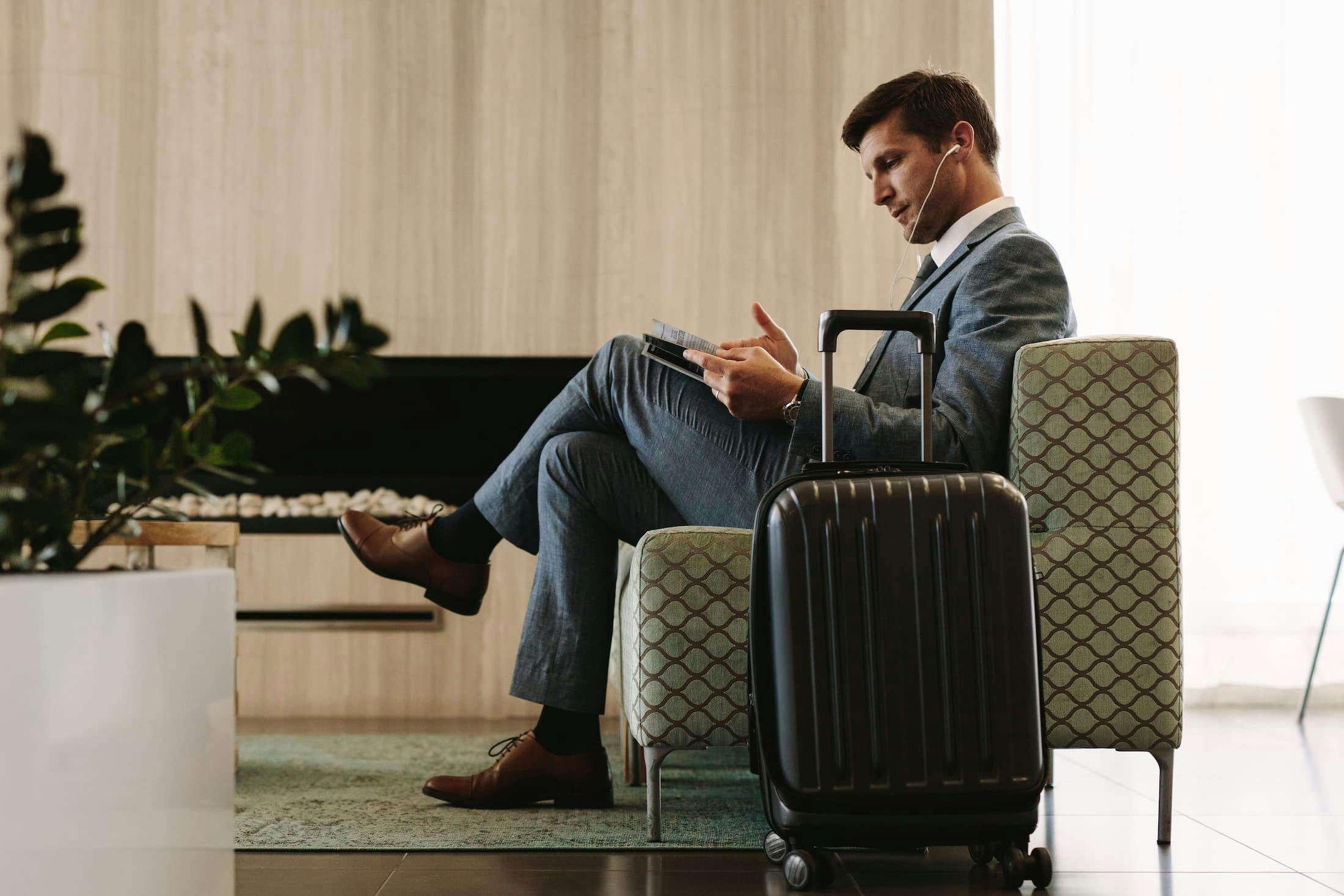Man in a suit sitting on a chair reading a magazine with a suitcase beside him.