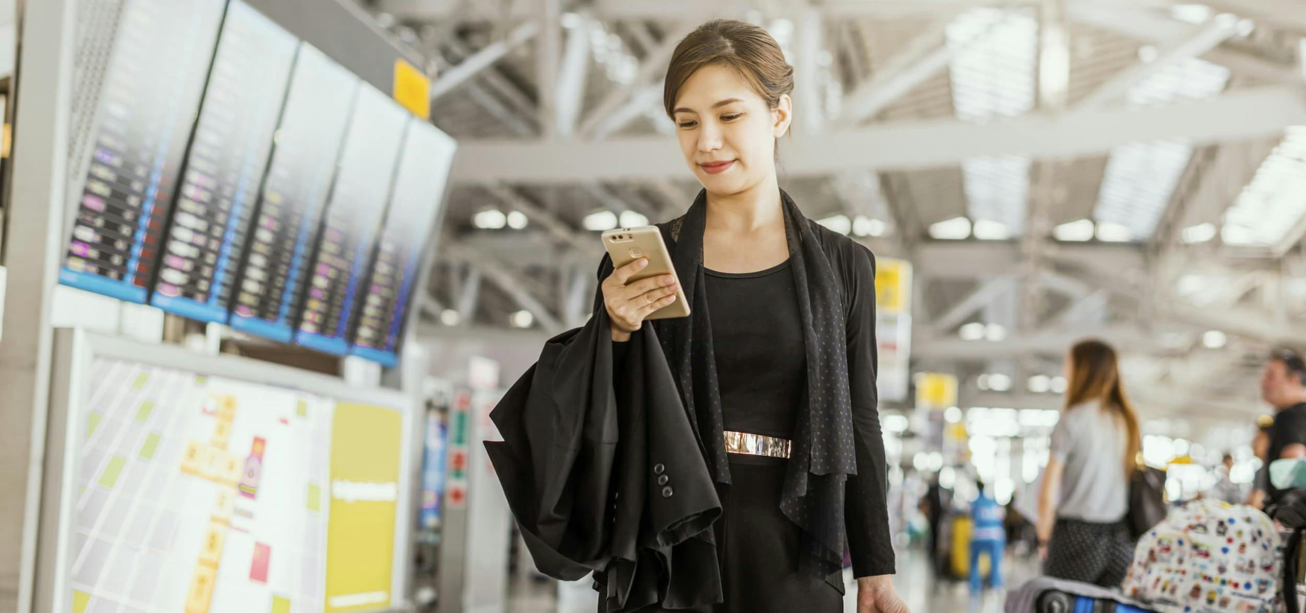 Woman in business clothes walking through an airport looking at her phone and holding a jacket.