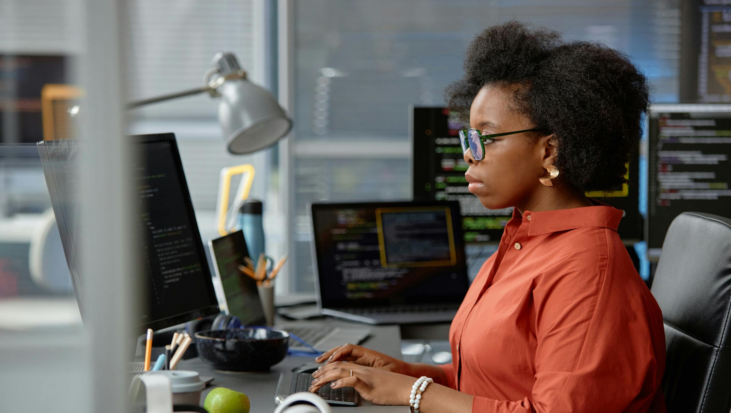 Side view shot of thoughtful female African American in glasses working on a computer with multiple screens.