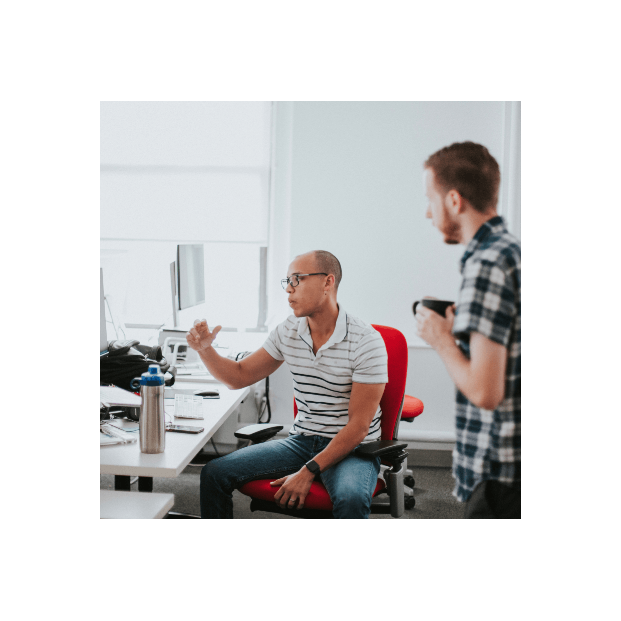 Man sitting at a desk talking about something he is working on while and other person looks on while holding a coffee mug.