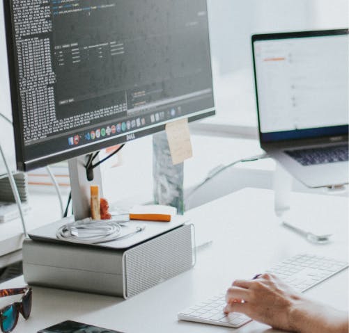 A monitor sitting on a book with code on the screen; below is a hand typing on a keyboard.