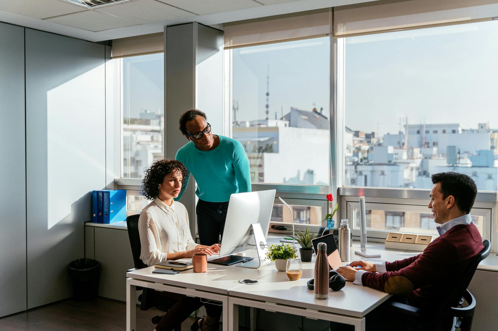3 people at a desk, one is a woman in a white shirt looking at a computer monitor with a man in a teal shirt looking over h er shoulder, across from them is a man in a burgundy sweater typing on a lapatop