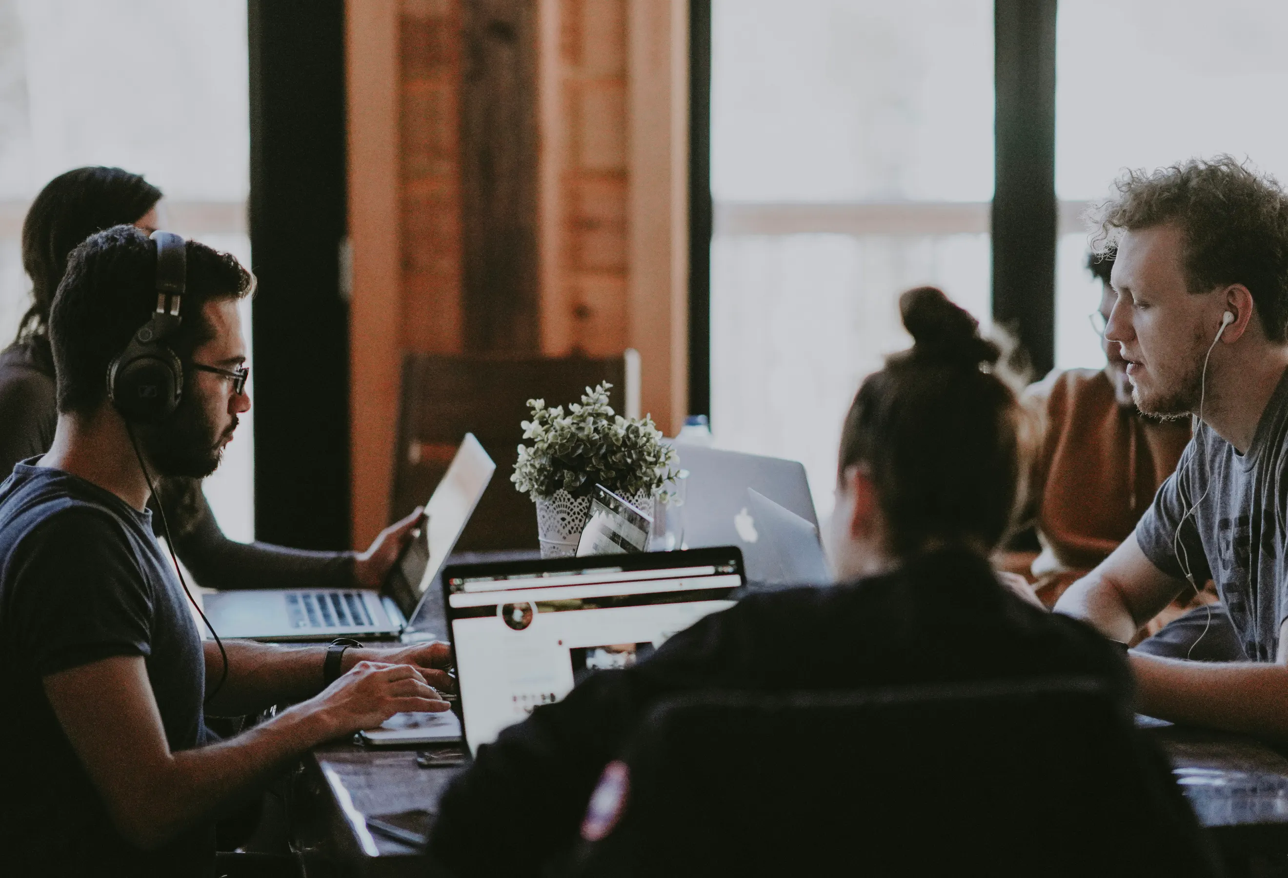 Various people sitting at a meeting table. Most people are wearing headphones and all are looking at their own laptops.