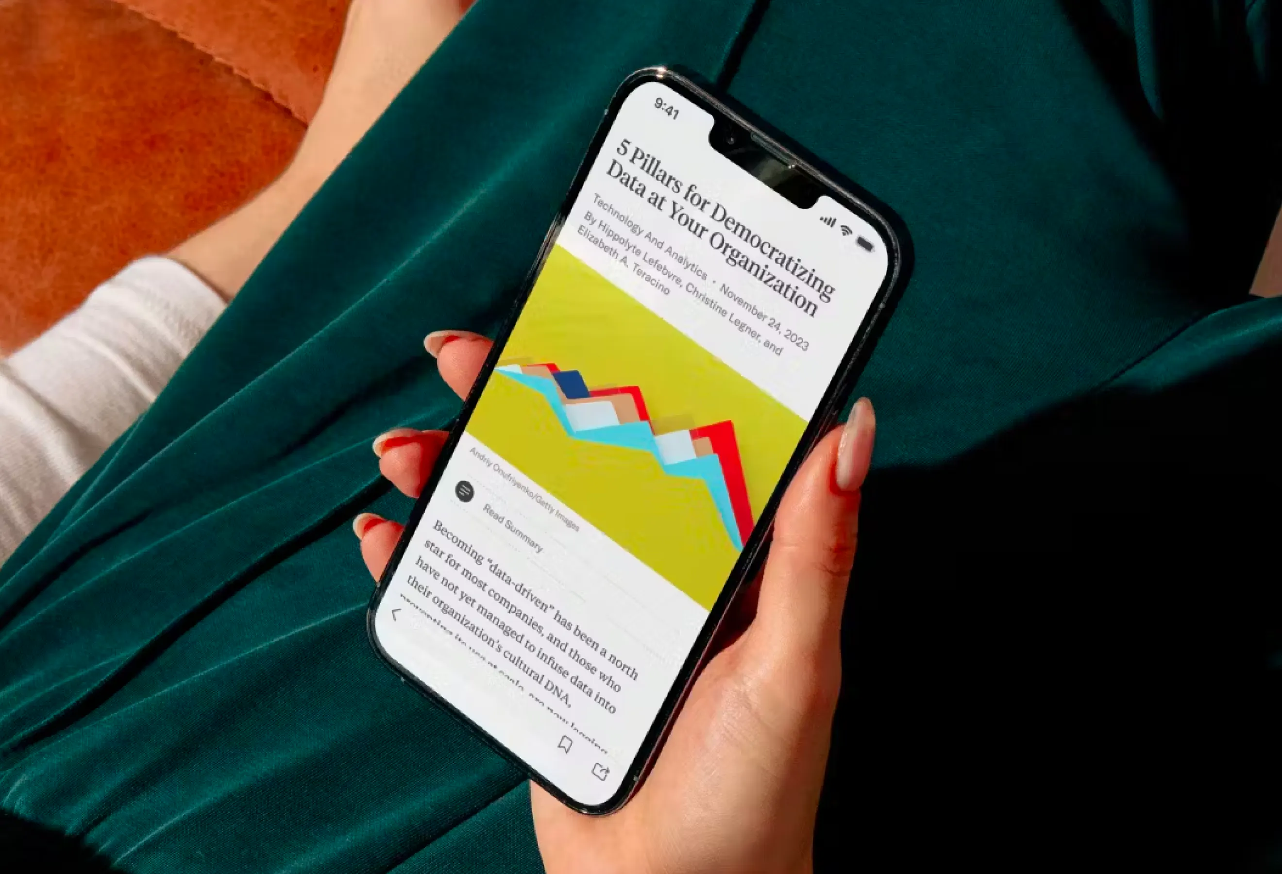 A cropped view of a person with green pants, focus is on their hand holding a phone. The screen has an article with an area chart image and a title that reads '5 Pillars for Democratizing Data at Your Organization'.
