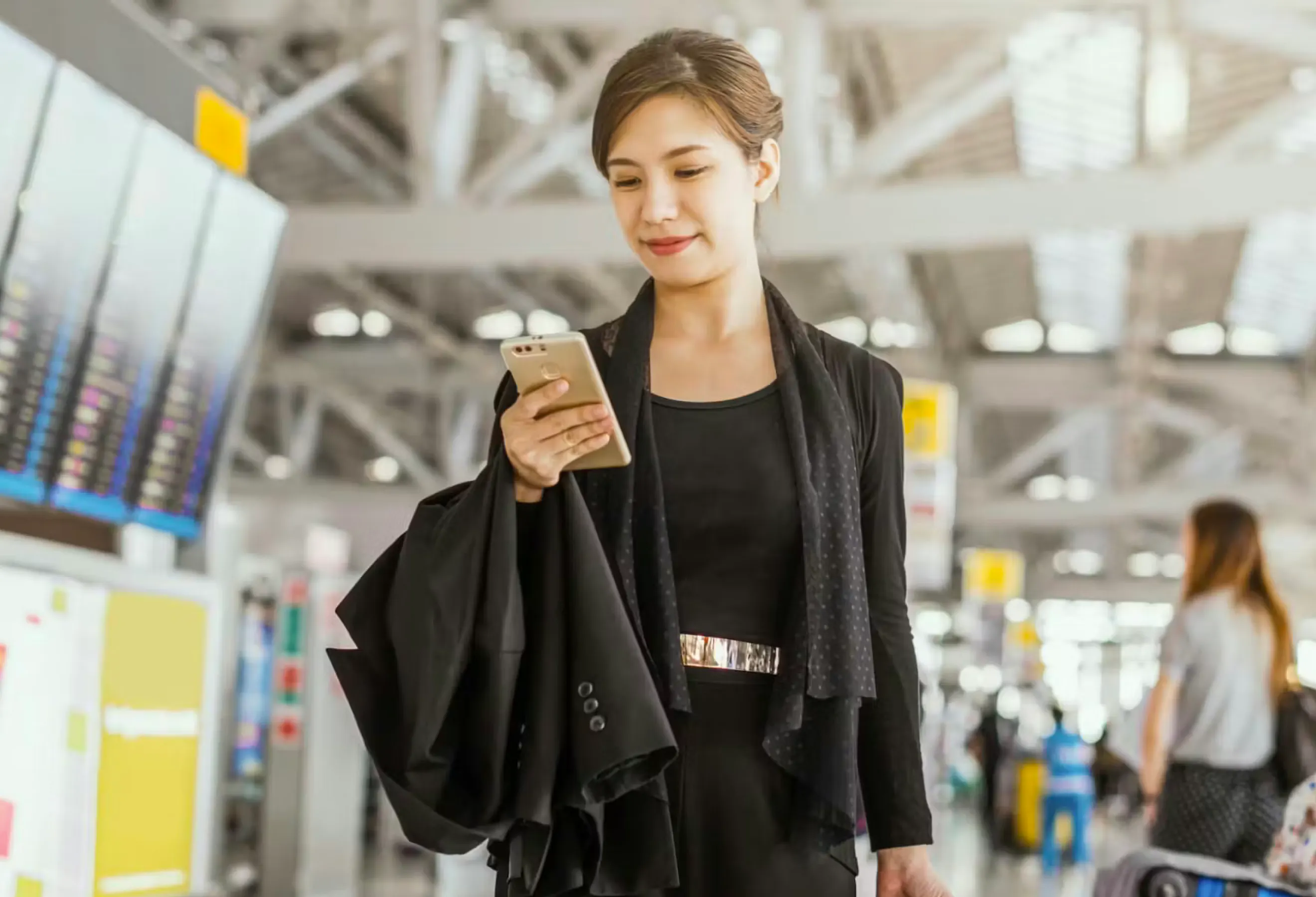 A woman wearing a black dress and shawl looking down at her phone while standing in an airport.