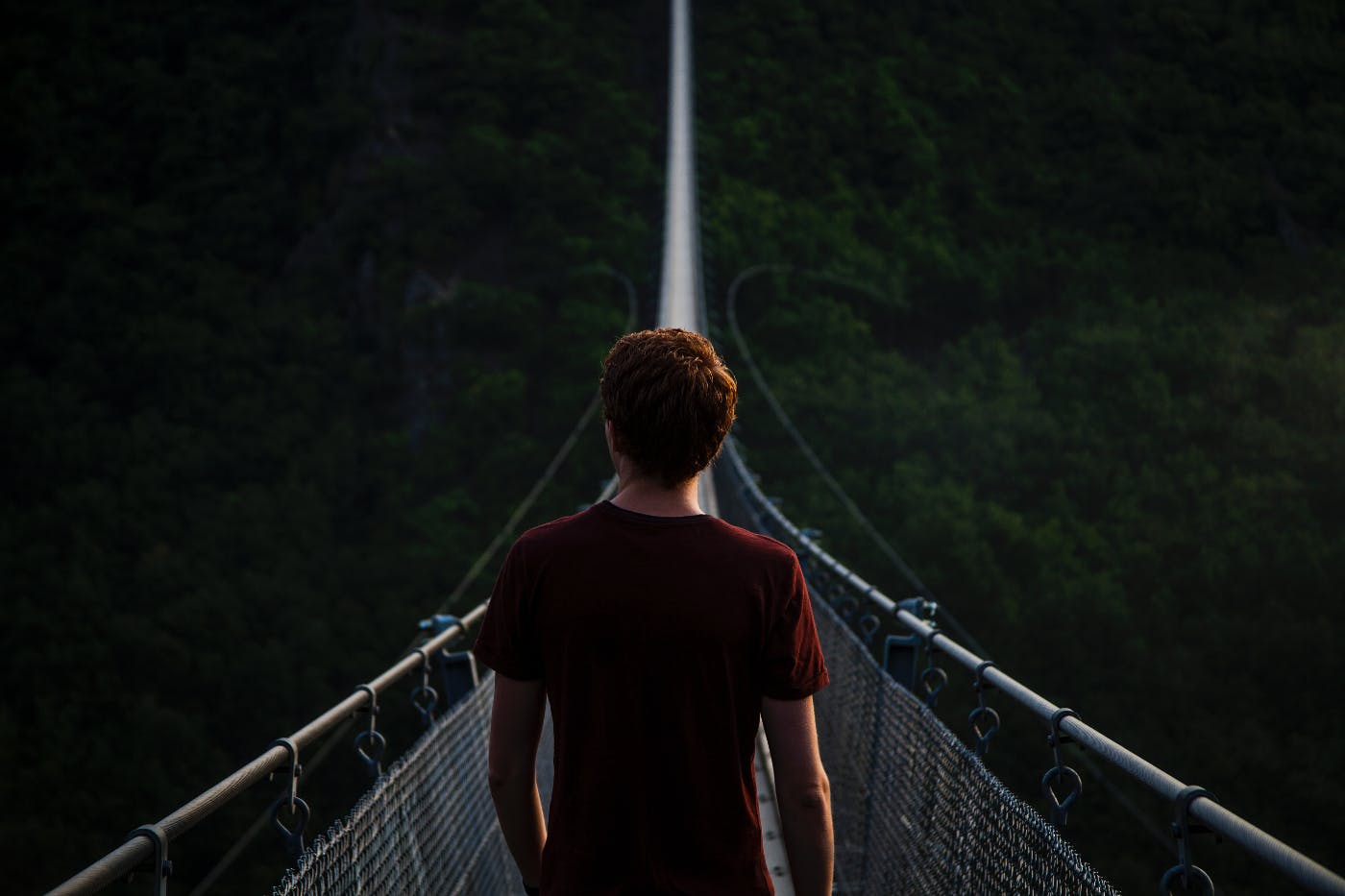 A young man standing at one end of a cable bridge stretched across a valley