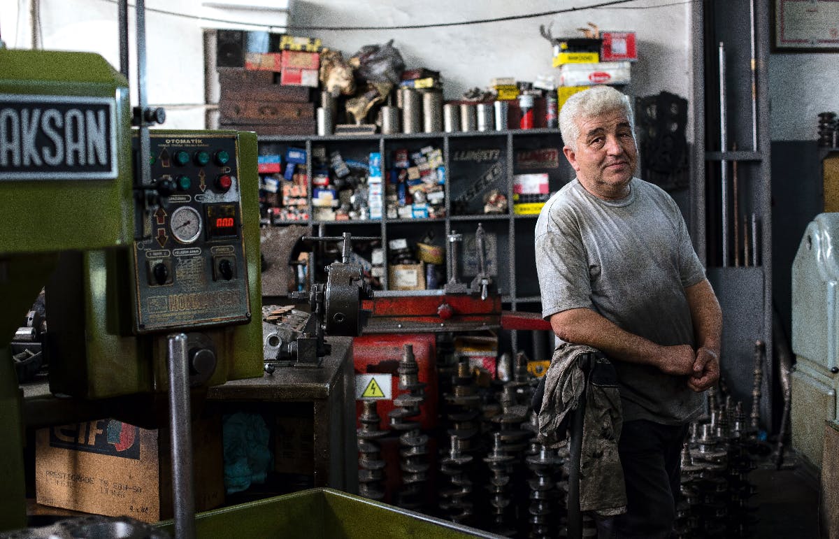 A man in a gre t-shirt in his work shop surrounded by mechanical gears and machines.