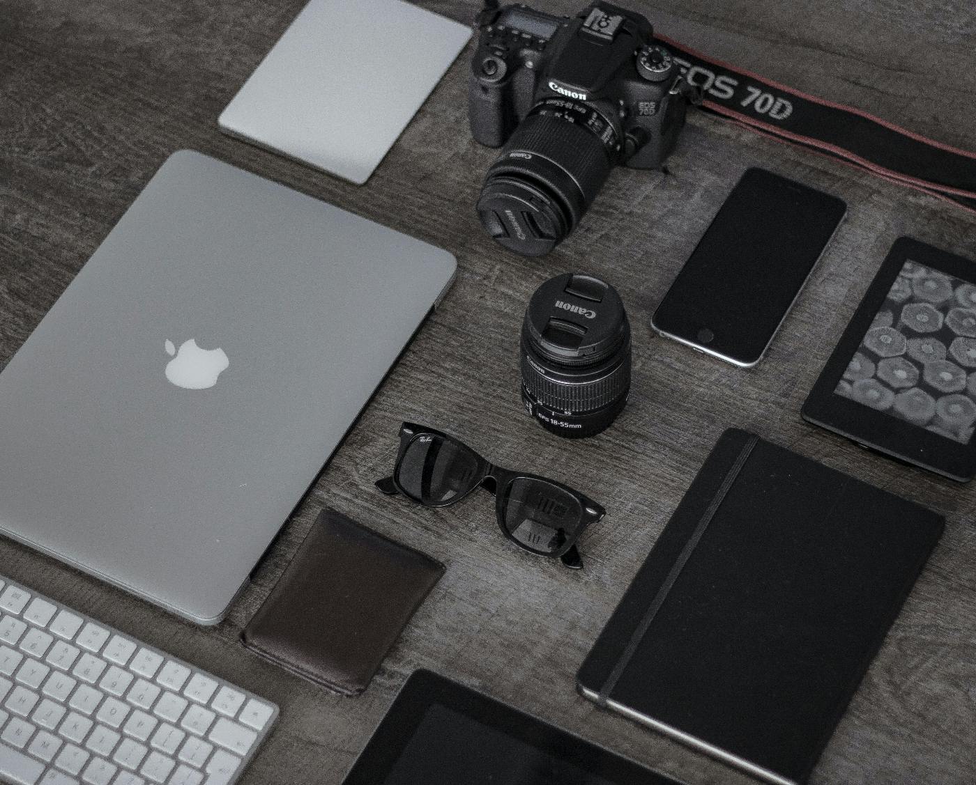 A table with a Macbook. Cannon camera and lens, iPhone, iPad, Raybans, moleskine and a wallet on it