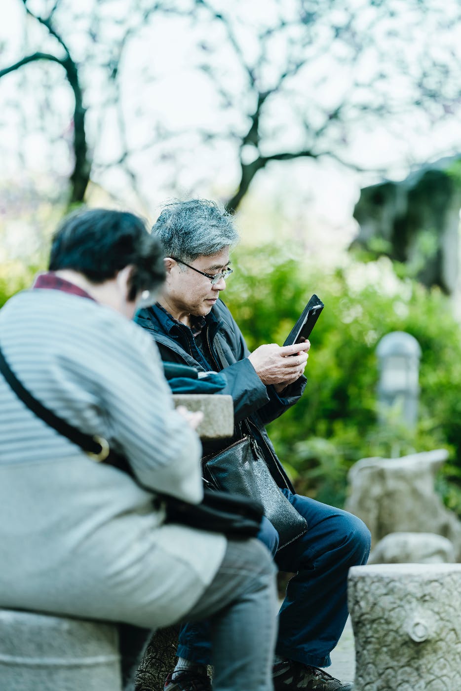 An older Chinese couple sitting on a stone bench looking at their smartphones