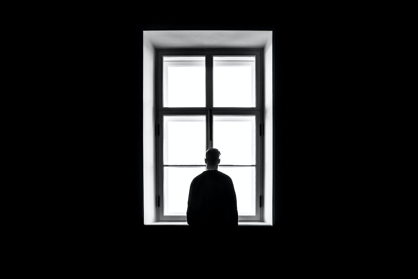 A man in a dark room standing looking out a window