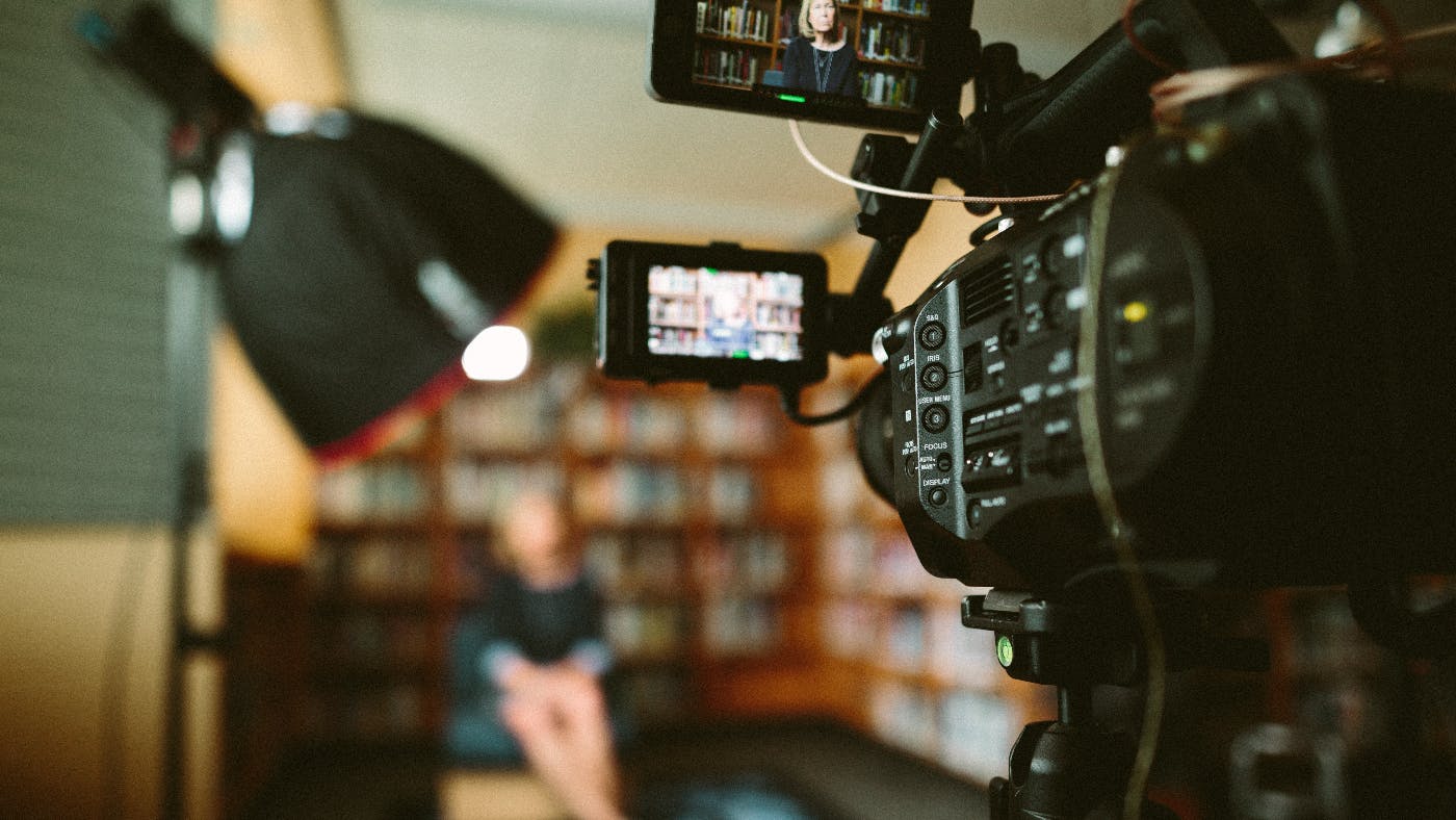 A video camera and light in the foreground, a woman sitting against bookshelves in the back