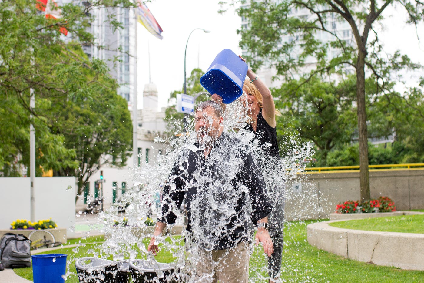 A woman pouring a bucket of ice water over a man's head in a park
