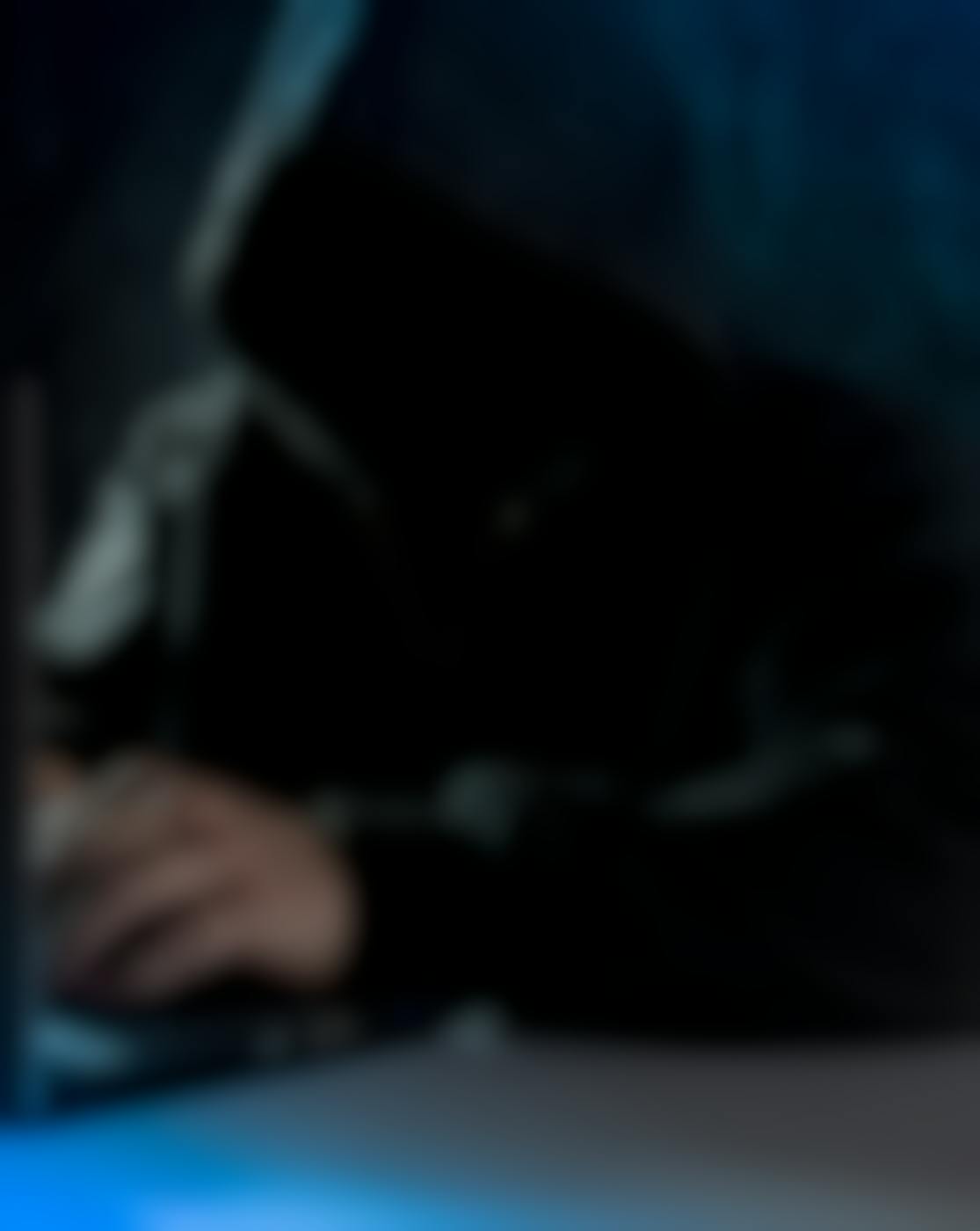 A faceless man in a black hoodie working on a laptop