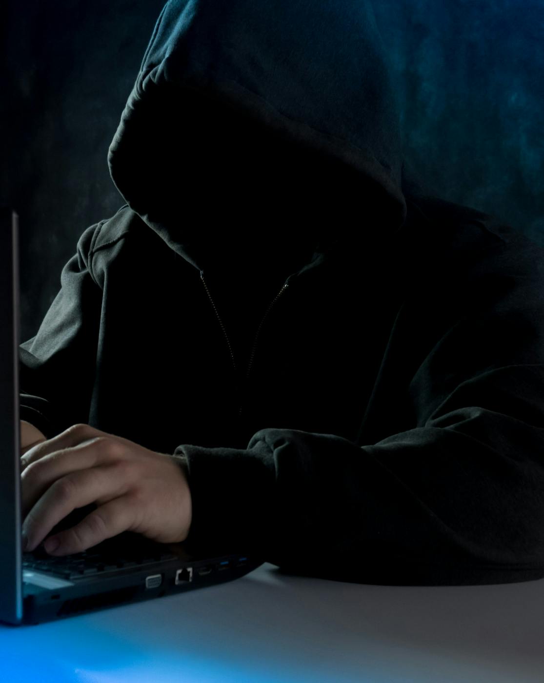 A faceless man in a black hoodie working on a laptop