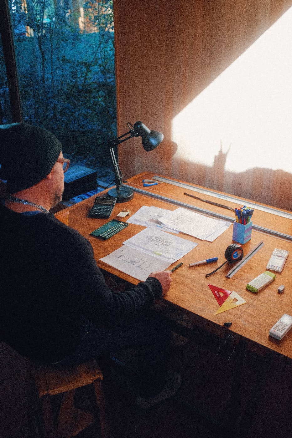 A designer in a black cap at his desk, design tools on the desk a desk lamp shines on the wall