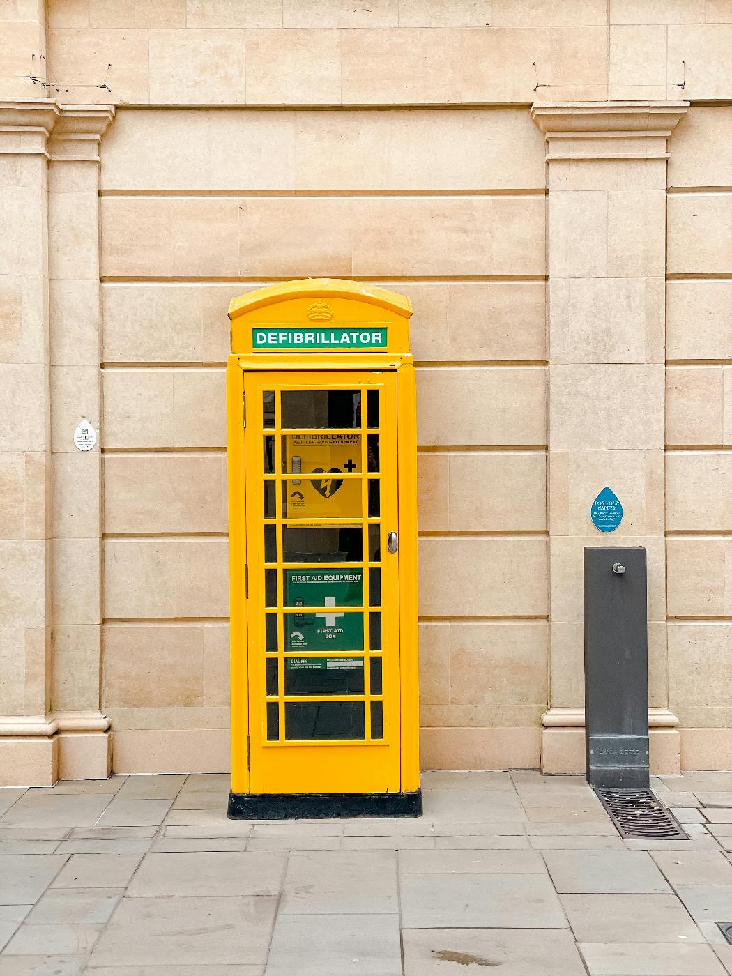 A yellow British style phone box with a green banner reading defibrillator