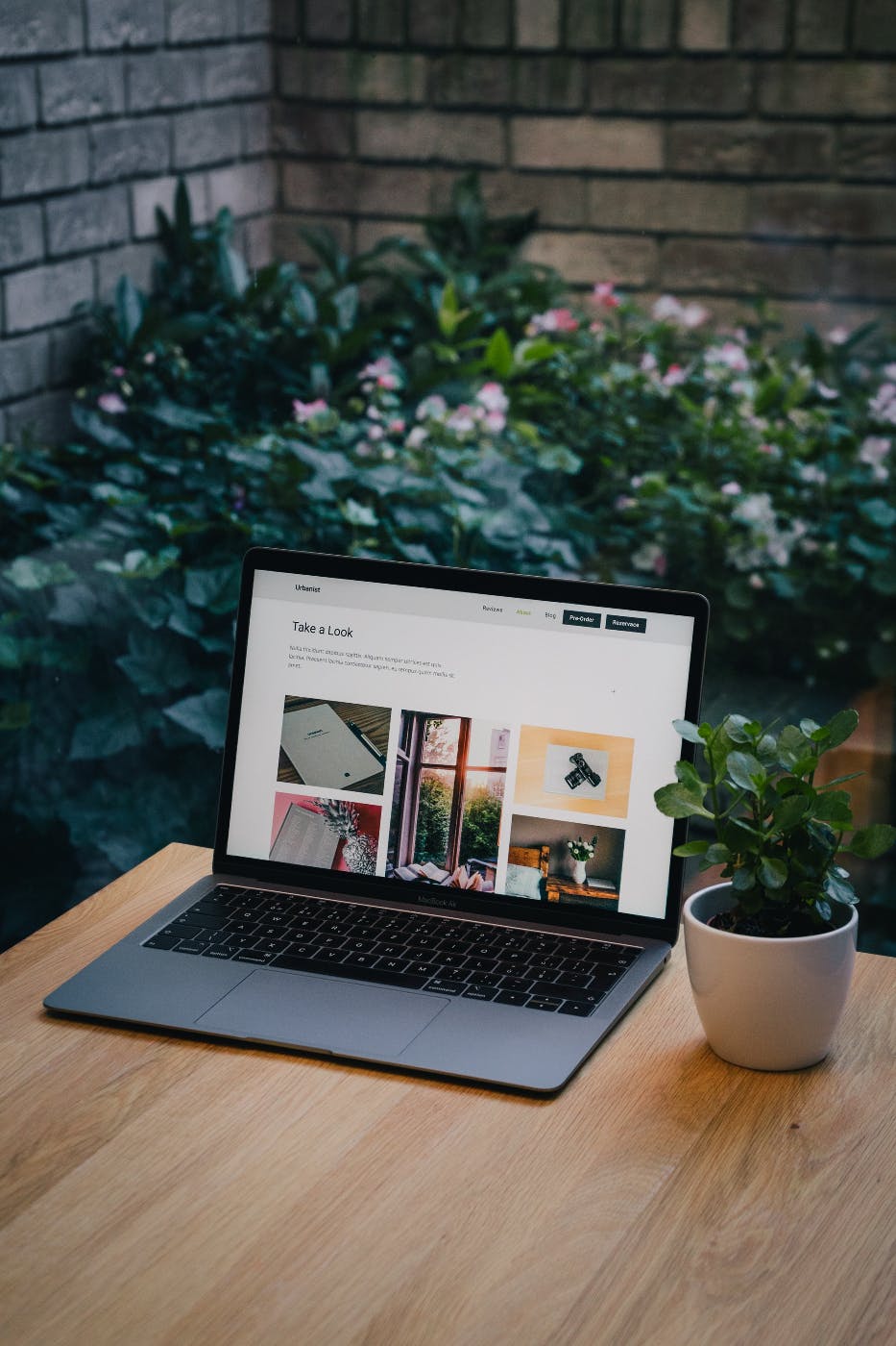 a lap top on a table next to a potted plant, in front of a brick wall and corner garden